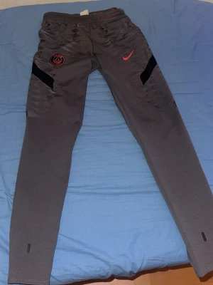 Grå PSG träningsbyxor från Nike - Snygga grå träningsbyxor från Nike med PSG-logga och röd Nike Swoosh på framsidan. Byxorna har svarta detaljer på låren och smal passform. Perfekta för fotbollsträning eller gymmet.