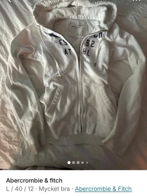 Vit hoodie från Abercrombie & Fitch - Vit zip-hoodie från Abercrombie & Fitch med tryck i mörkblått på bröstet och snörning i huvan. Mjuk bomullskänsla, två fickor fram och klassisk passform. Perfekt för en avslappnad och stilren look. Skulle säga att den sitter mer som en storlek S eller M snarare än L. 