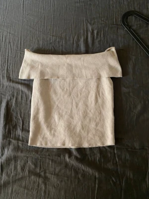 Beige offshoulder topp från H&M - Säljer en beige offshoulder topp från H&M i mjukt stickat material. Toppen är axelbandslös och har en bred kant upptill som ger en snygg och trendig look. Perfekt att styla med jeans eller kjol för en cool outfit.