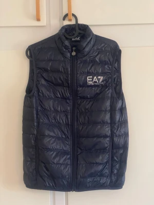 Mörkblå dunväst från EA7 Emporio Armani - Snygg mörkblå dunväst från EA7 Emporio Armani med quiltad design och hög krage. Västen har dragkedja framtill och logga på bröstet. Perfekt för lager-på-lager och streetstyle. Materialet är lätt och ger en sportig look.
