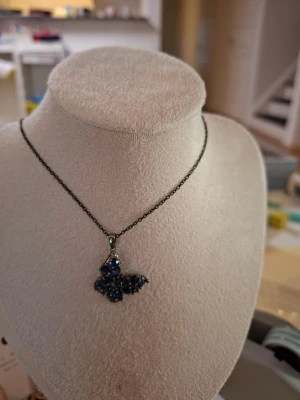 Halsband med blå fjärilspendel - Snyggt halsband med en fjärilsformad berlock dekorerad med blå pärlor och stenar. Kedjan är mörk och metallisk, vilket ger en cool kontrast till de glittrande blå detaljerna. Perfekt för dig som gillar unika smycken med lite extra färg.