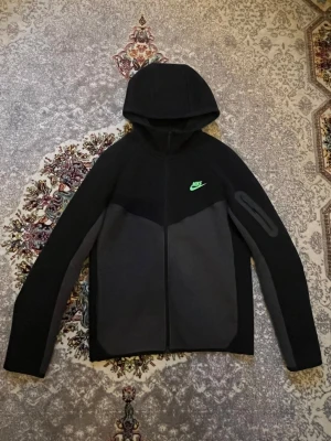 Nike Tech Fleece  - Nike Tech Fleece hoodie i svart och mörkgrått med hel dragkedja, huva och en grön Nike-logga på bröstet. Snygg detalj med ficka med dragkedja på ärmen. Tillverkad i mjukt och stretchigt fleecematerial som är perfekt för en sportig och avslappnad stil. Pris kan diskuteras!