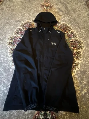 Svart vindjacka från Under Armour - Snygg svart vindjacka från Under Armour med huva och dragkedja framtill. Jackan har reflekterande detaljer på axlarna och logga på bröstet. Tillverkad i lätt och tåligt material, perfekt för blåsiga dagar. Justerbar huva med snören och fickor på jackan. Zippen är lite krånglig annars inga probelm