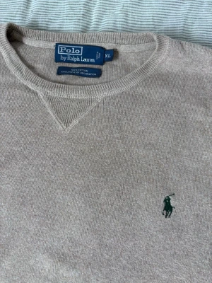Ralph Lauren knit - Snygg beige stickad tröja från Ralph Lauren i bomull. Klassisk rund halsringning och lång ärm, med den ikoniska gröna loggan broderad på bröstet. Sitter oversized i XL