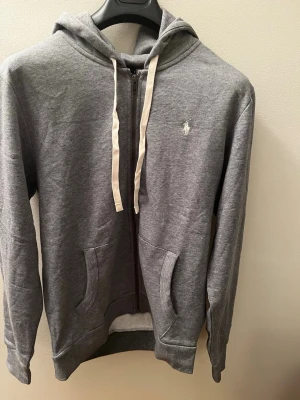 Grå hoodie från Polo Ralph Lauren - Snygg grå zip-hoodie från Polo Ralph Lauren med vit logga på bröstet. Hoodien har huva med vita snören, två fickor fram och dragkedja. Perfekt för en chill och avslappnad stil.