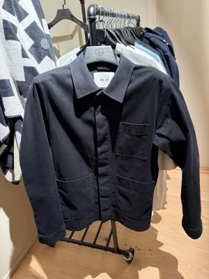 Svart overshirt från NN.07 - Stilren svart overshirt från NN.07 med klassisk krage och tre praktiska fickor framtill. Overshirten är tillverkad i ett mjukt och slitstarkt tyg, perfekt för lager-på-lager och en clean look.