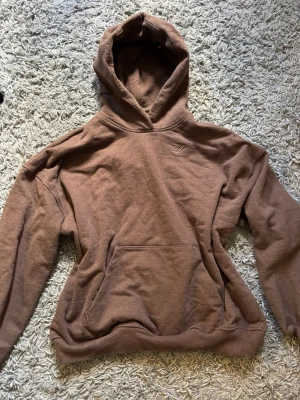 Brun hoodie från Gymshark - Säljer en brun hoodie från Gymshark med broderad logga på bröstet. Hoodien har en stor känguruficka framtill, ribbade muddar och en rejäl huva. Den är i stolek S. 