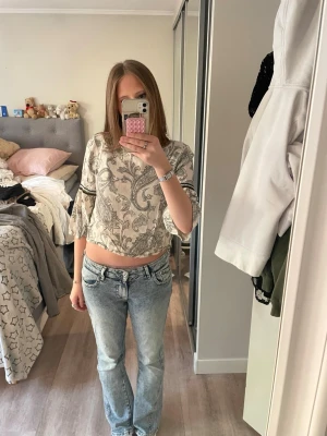 Beige croppad blus med paisleymönster - Säljer en beige croppad blus med svart paisleymönster och vida trekvartsärmar. Toppen har dekorativa banddetaljer på ärmarna och är tillverkad i ett mjukt, tunt material som känns skönt mot huden. Perfekt för dig som gillar boho-vibbar och unika detaljer.