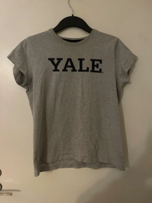 Grå t-shirt med YALE-tryck - Snygg grå t-shirt med svart YALE-tryck på bröstet. Klassisk passform med rund hals och korta ärmar. Perfekt för en avslappnad och cool stil. Tillverkad i mjuk bomull som känns skön mot huden.