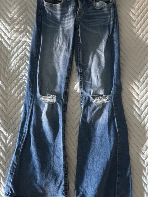 Blå bootcut jeans med slitningar - Säljer ett par blå bootcut jeans med slitningar över knäna och coola röda sömmar på bakfickan. Jeansen har klassisk femficksmodell och är i mjukt denimtyg. Perfekta för en avslappnad och trendig look.