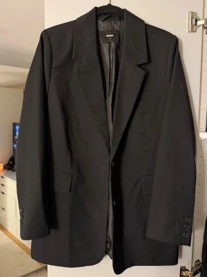Svart kavaj från Bikbok - Stilren svart kavaj från Bikbok med klassisk passform, två knappar framtill och fickor med lock. Kavajen har breda slag och är helfodrad för en snygg siluett. Perfekt för att lyfta vilken outfit som helst.