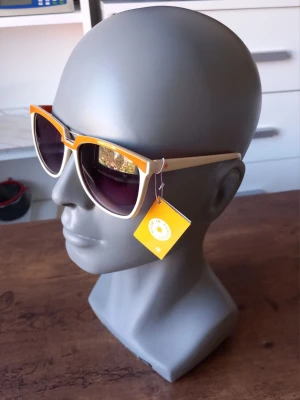 Vita och orangea solglasögon från Sunshine  - Snygga solglasögon från Sunshine med vit båge och orange detaljer. Modellen har en klassisk form med breda skalmar och mörka glas. Perfekta för att sticka ut och ge din look en färgklick. Passar dig som gillar retrovibbar och coola accessoarer.