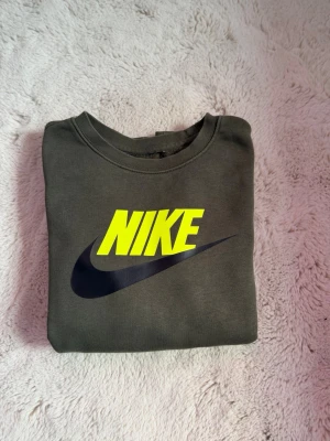 Nike sweatshirt med neonlogga - Mörkgrön sweatshirt från Nike med stor gul neonlogga och svart swoosh på bröstet. Tröjan har rund halsringning, långa ärmar och ribbade muddar. Perfekt för en sportig och avslappnad stil.