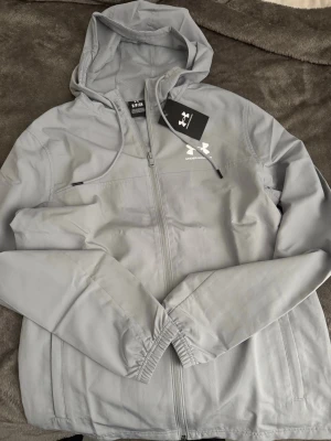 Ljusgrå vindjacka från Under Armour - Snygg ljusgrå vindjacka från Under Armour med huva och dragkedja framtill. Jackan har elastiska muddar vid ärmsluten, två sidofickor och en vit logga på bröstet. Perfekt för blåsiga dagar och sportiga outfits Jackan är helt ny, Aldrig använd. Storlek S UA Rival.