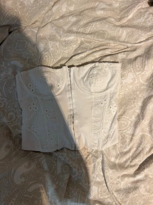Vit korsett från Zara med broderi - Supersnygg vit korsett från Zara med broderade detaljer och markerade kupor. Toppen är axelbandslös och har fina hålbroderier som ger en romantisk vibe. Perfekt att styla med jeans eller kjol för en trendig look.