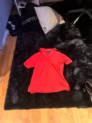 Röd piké från Ralph Lauren - Snygg röd pikétröja från Ralph Lauren med klassisk krage och knappar framtill. Tröjan har korta ärmar och den ikoniska blå loggan broderad nertill. Perfekt till jeans eller shorts för en avslappnad look.