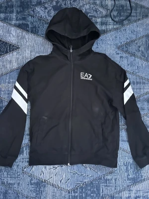 Svart EA7 hoodie med vita ränder - Svart hoodie från EA7 Emporio Armani med dragkedja, huva och vita ränder på ärmarna. Logga på bröstet och ryggen. Perfekt för en sportig och stilren look. Mjukt material och två fickor framtill.
