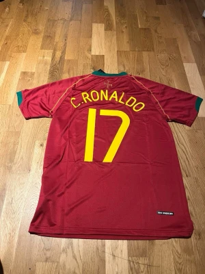 Portugal Ronaldo 17 matchtröja Nike - Röd Portugal fotbollströja från Nike med gult nummer 17 och C. Ronaldo på ryggen. Tröjan har gröna detaljer vid krage och ärmslut samt Portugals landslagsemblem på bröstet. Perfekt för dig som älskar fotboll och vill hylla Ronaldo.