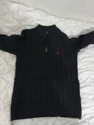 Svart kabelstickad tröja Polo Ralph Lauren - Svart kabelstickad tröja från Polo Ralph Lauren med halv dragkedja och röd broderad logga på bröstet. Tröjan har ribbade muddar och hög krage, perfekt för lager-på-lager-stil.  Pris kan diskuteras priv 