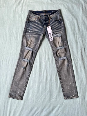 Grå slitna jeans från Purple Brand - Säljer ett par grå jeans från Purple Brand med coola slitningar och hål på benen. Jeansen har vit färgstänk och en unik tvättad look med ränder vid fickorna. Klassisk femficksmodell och skinny fit. Perfekta för en edgy streetstyle.