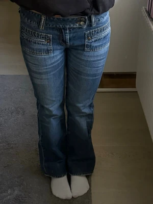 Blå bootcut jeans med fickdetaljer - Säljer ett par blå bootcut jeans med snygga fickdetaljer framtill och låg midja. Jeansen har slitningar och kontrastsömmar som ger en cool vintagekänsla. Perfekta för dig som gillar retrostil och vill ha ett par unika jeans i garderoben.