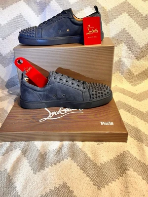 Christian Louboutin mörk grå sneakers - Mörkgråa sneakers i mocka från Christian Louboutin med ikoniska röda sulor och gråa nitar på tån. Lågt skaft, grå skosnören och diskreta metalldetaljer. Perfekta för dig som vill sticka ut med en edgy och lyxig stil. (Allt på bilderna medföljer)