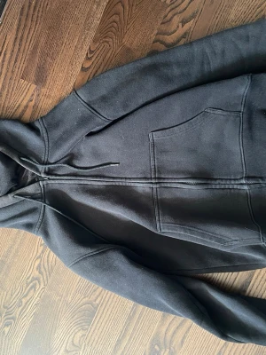 Svart hoodie med grafiskt tryck - Svart hoodie med dragkedja och huva, samt stora fickor framtill. Baksidan har ett unikt vitt grafiskt tryck i form av ett cirkelmönster. Perfekt för dig som gillar streetwear och vill sticka ut lite extra. Mjuk och skön bomullsblandning.