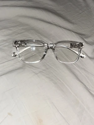 Chrome heart glasögon - Unika solglasögon med transparent båge och rektangulära glas. Snygga dödskalle-detaljer i metall på sidorna ger en edgy vibe. Perfekta för dig som vill sticka ut med en cool accessoar. Materialet är plast och metall.