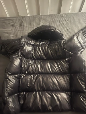 Svart glansig dunjacka med huva - Snygg svart dunjacka med glansig finish och stor huva. Jackan har breda kanaler, dragkedja framtill och två sidofickor med dragkedja. Tillverkad av återvunnet material, perfekt för kalla vinterdagar, jackan är knappt använd och jättefräsch. .