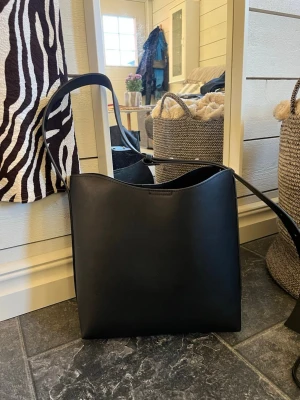 Svart axelväska i minimalistisk design - Stilren svart axelväska i slät konstläder med enkel, fyrkantig form och lång axelrem. Perfekt för dig som gillar en clean och modern look. Väskan har inga synliga loggor eller detaljer, vilket gör den lätt att matcha med olika outfits.
