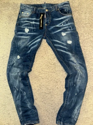 Dsquared2 blå jeans - Säljer ett par blå jeans från Dsquared2 med slitna detaljer, färgstänk och dragkedja framtill. Jeansen har en smal passform och klassisk femficksdesign. De är äkta!! Fraktas samma dag eller dagen efter
