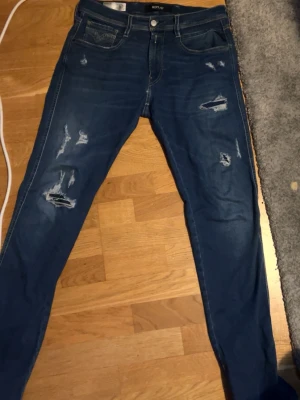 Replay jeans - Säljer ett par blå slim fit jeans från Replay, modell Anbass. Jeansen har slitna detaljer och hål på benen för en trendig look. Klassisk femficksmodell med knapp och dragkedja. Perfekta för dig som gillar en modern och avslappnad stil.