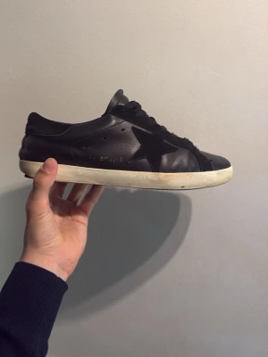 Svarta Golden Goose sneakers i Italien läder - Svarta sneakers från Golden Goose i läder med ikonisk stjärndetalj på sidan och svarta skosnören. Skon har en vit, något sliten sula och klassisk låg profil. Inga riktigt skönhets problem förutom sulan. Är riktigt sköna och pris kan diskuteras  