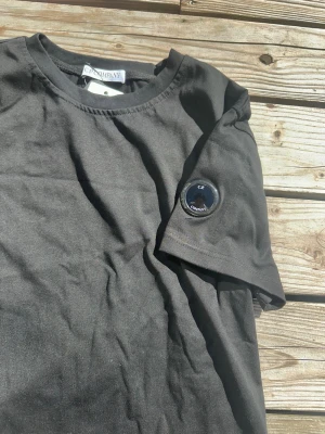 Svart t-shirt från C.P. Company - Snygg svart t-shirt från C.P. Company med klassisk rund hals och korta ärmar. På ena ärmen finns det ikoniska linsen med loggan. Tillverkad i mjuk bomull som känns skön mot huden. Perfekt för en clean och stilren look