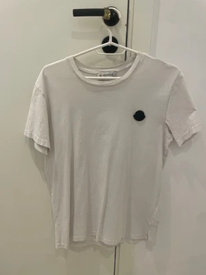 Vit Moncler t-shirt med svart logga - Vit t-shirt från Moncler med svart logga på bröstet och stor vertikal Moncler-text på ryggen. Klassisk rund halsringning och korta ärmar. Perfekt för en clean och stilren look.