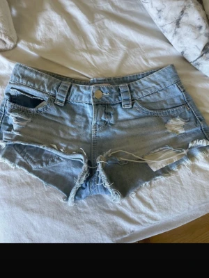 Ljusblåa slitna jeansshorts - Säljer ett par ljusblåa jeansshorts med slitna detaljer och fransig kant. Skulle säga att de passar en tjej med storlek S med! Shortsen har klassisk femficksdesign, bälteshällor och knappstängning framtill. Perfekta för varma dagar och en avslappnad stil.