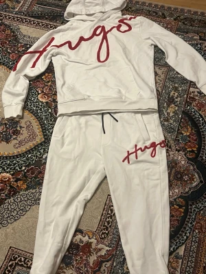 Vit loungewear-set från Hugo - Snyggt vitt loungewear-set från Hugo med röd logga i stor, handskriven stil på både hoodie och byxor. Hoodien har magficka och byxorna har dragsko i midjan. Perfekt för chill dagar hemma eller när du vill vara bekväm men ändå se fräsch ut.