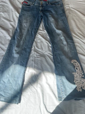 Blå bootcut jeans med spetsdetalj - Säljer ett par ljusblå bootcut jeans med snygg vit spetsapplikation på ena benet. Jeansen har klassisk femficksmodell och normal midja. Perfekta för dig som vill ha en unik twist på dina denim! Passar till sneakers eller boots.
