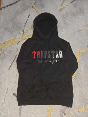Svart hoodie från Trapstar med tryck - Svart hoodie från Trapstar med färgglatt tryck på bröstet i rött, grått och vitt. Klassisk huva med dragsko och magficka framtill. Mjuk insida och ribbade muddar vid ärmslut och nederkant. Perfekt för en streetwear-look.