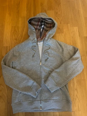 Grå zip-hoodie från Burberry - Snygg grå hoodie med dragkedja från Burberry. Klassisk huva med det ikoniska rutmönstret på insidan och broderad logga på bröstet. Mjukt material och ribbade muddar. Perfekt för en chill och stilren look.