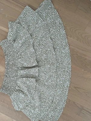 Grön blommig kjol från SHEIN - Säljer en grön kortkjol från SHEIN med vitt blommönster. Kjolen har resår i midjan och är gjord i ett lätt och luftigt bomullstyg. Perfekt för dig som gillar en avslappnad och söt stil.