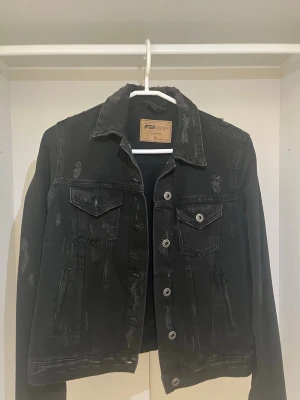 Svart jeansjacka från FB Sister - Cool svart jeansjacka från FB Sister i storlek S. Jackan har slitna detaljer, klassisk krage, två bröstfickor med lock och knappar framtill. Perfekt för dig som gillar en edgy och trendig stil.