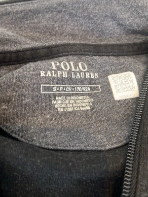 Svart halvzip från Polo Ralph Lauren - Klassisk svart långärmad tröja från Polo Ralph Lauren med halv dragkedja och diskret logga på bröstet. Tillverkad i mjukt material och har ribbade muddar vid ärmslut och nederkant. Perfekt för en stilren och avslappnad look.