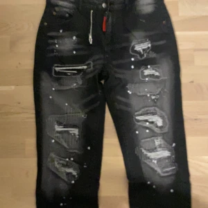 Svarta jeans med slitningar och färgstänk  - Svarta jeans med kraftiga slitningar och vita färgstänk framtill. Jeansen har en modern, edgy look med detaljerade trasiga partier och kontrasterande ljusa partier. Passar dig som vill sticka ut med din stil. Materialet är en blandning av bomull och syntet. Ser också ut som dsquard2 Jeans men tyvär är dem inte de 