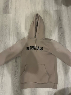 Beige Essentials Fear of God hoodie - Snygg beige hoodie från Fear of God Essentials med svart tryck på bröstet och ärmen. Klassisk känguruficka framtill och stor huva. Tillverkad i mjukt bomullsmaterial som känns riktigt skönt mot huden. Perfekt för dig som gillar streetwear och enkel stil.