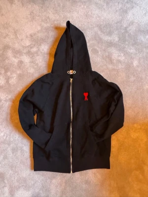 Ami paris hoodie - Svart hoodie med dragkedja från Ami Paris. Klassisk modell med huva och snörning, samt en liten röd Ami-hjärta-logga broderad på bröstet. Tillverkad i mjukt bomullsmaterial och har fickor framtill. Ny pris 4450kr.