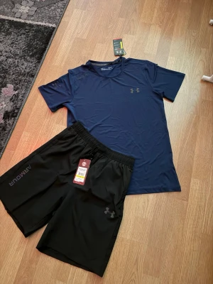 Under Armour träningsset blå/svart storlek M - Snyggt träningsset från Under Armour med en mörkblå t-shirt i funktionsmaterial och svarta shorts med elastisk midja. Perfekt för gymmet eller löprundan. Båda plaggen har diskreta loggor och är lätta och bekväma.