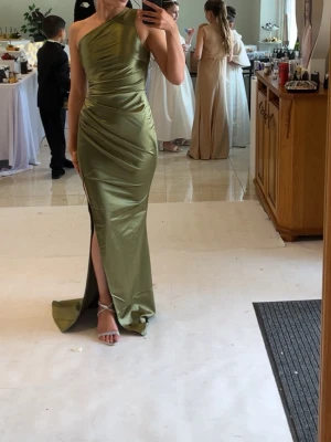 Grön one shoulder långklänning - Snygg grön långklänning i satin med one shoulder-design och hög slits framtill. Klänningen har figurnära passform och draperade detaljer som ger en elegant siluett. Perfekt för speciella tillfällen.