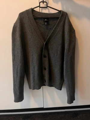 Mörkgrå kofta från H&M - Mysig mörkgrå kofta från H&M med v-ringning och knäppning framtill. Koftan har långa ärmar och ribbade muddar vid ärmslut och nederkant. Perfekt att slänga på sig när det är lite kyligt ute.
