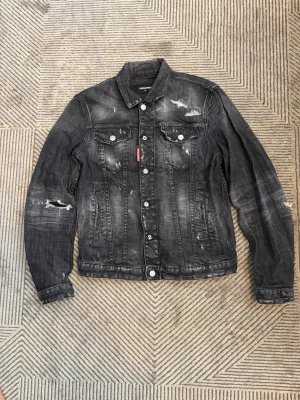 Svart distressed jeansjacka Dsquared2 - Cool svart jeansjacka från Dsquared2 med slitna detaljer och snygg tvätt. Klassisk modell med bröstfickor, knappar med Dsquared2-logga och röd logotag på bröstet. Perfekt för dig som gillar streetstyle och vill sticka ut.